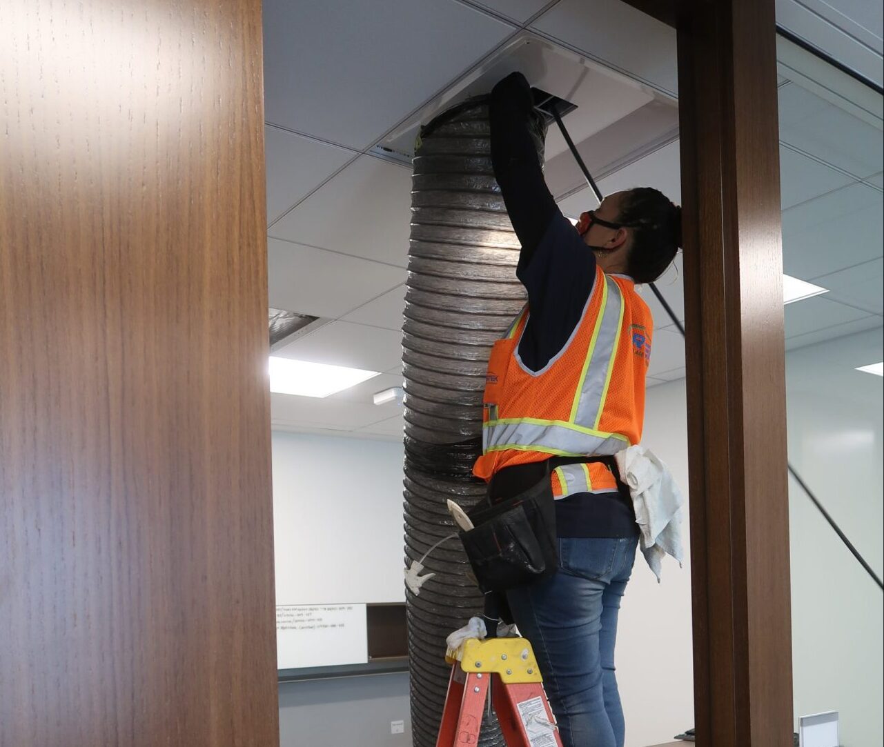 Worker in a safety vest standing on a ladder while performing duct cleaning in an office ceiling.